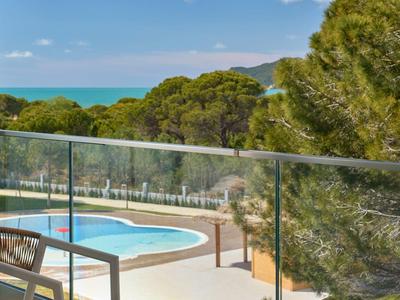 Blick auf Pool mit Glasgeländer, zwei Stühlen und Tisch, umgeben von Bäumen und blauem Himmel.