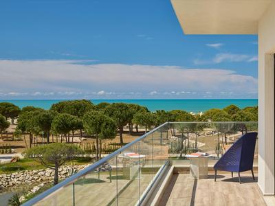 Balkon mit Glasgeländer, Blick auf grüne Bäume und blaues Meer unter klarem Himmel.