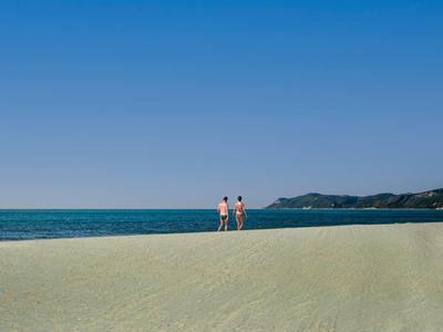 Zwei Personen stehen am Sandstrand mit klarem blauen Himmel und Meer im Hintergrund.
