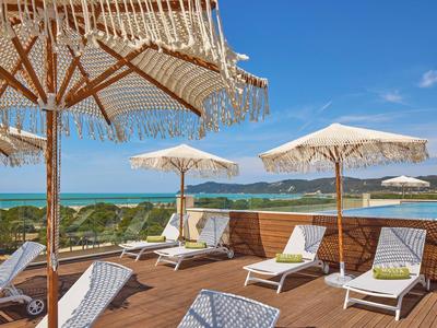 Terrassenbereich mit Holzdecke, weißen Liegestühlen und Sonnenschirmen vor blauem Himmel und Meer.