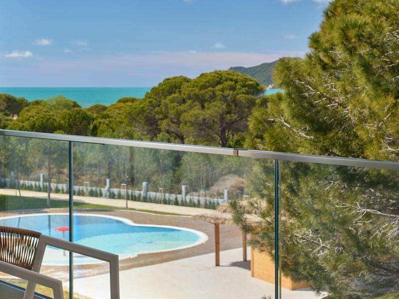 Blick auf Pool mit Glasgeländer, zwei Stühlen und Tisch, umgeben von Bäumen und blauem Himmel.