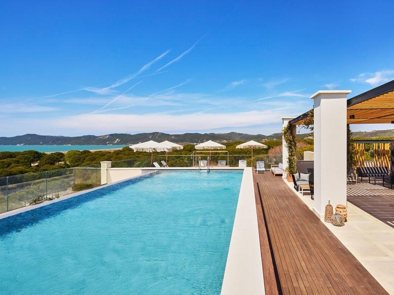 Moderner Pool mit Glasgeländer, Holzterrasse und Loungemöbeln, dahinter grüne Hügel und blauer Himmel.