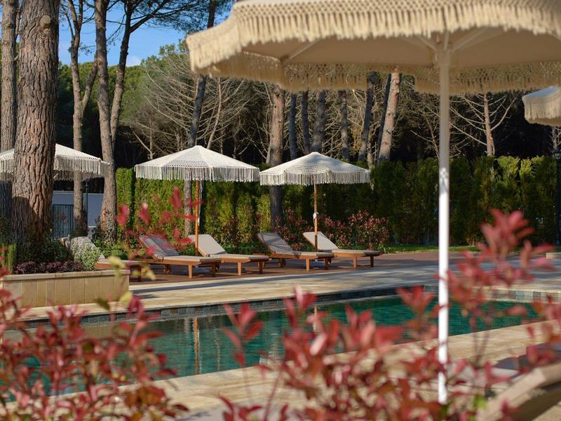 Poolbereich mit Sonnenliegen und großen, cremefarbenen Sonnenschirmen in einer bewaldeten Umgebung.