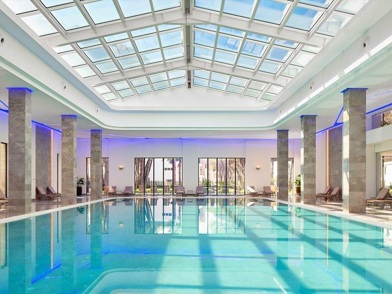 Helles Hallenbad mit Glasdach, Säulen, blauer Beleuchtung und Liegestühlen am Rand.