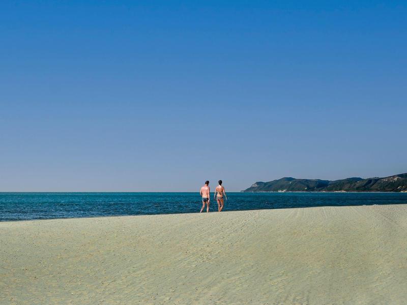 Zwei Personen stehen am Sandstrand mit klarem blauen Himmel und Meer im Hintergrund.