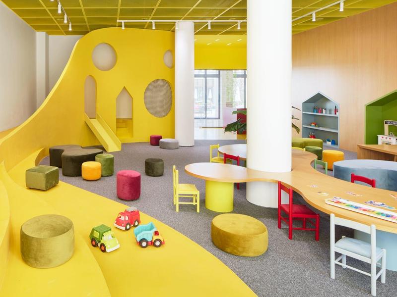 Bunter moderner Kindergartenraum mit gelber Rutsche, bunten Sitzmöbeln und Spielzeugautos.
