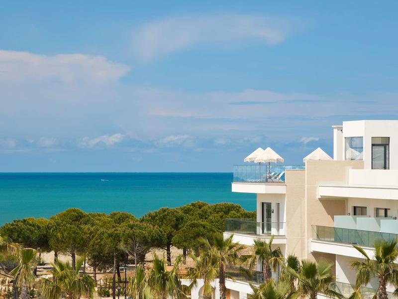 Blick auf blaues Meer, grünen Wald und weiße moderne Gebäude unter klarem Himmel.