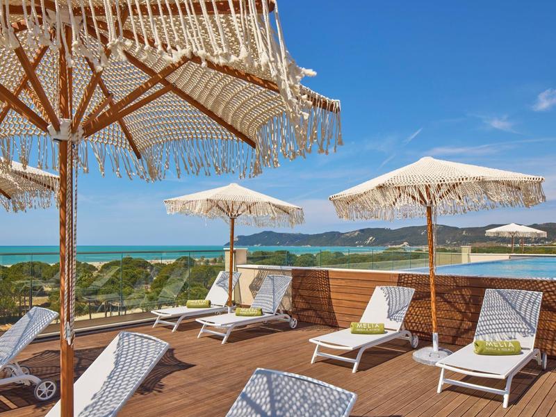 Terrassenbereich mit Holzdecke, weißen Liegestühlen und Sonnenschirmen vor blauem Himmel und Meer.