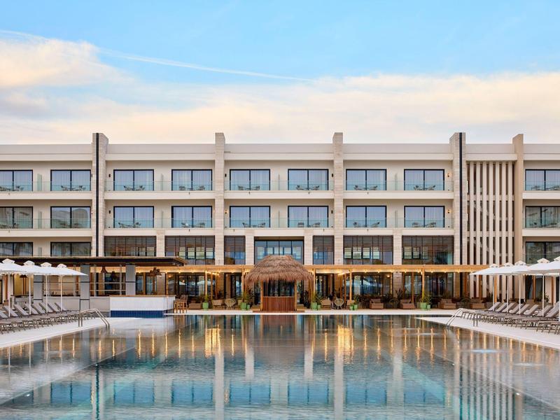 Moderner Hotelkomplex mit rechteckigem Pool und weißen Liegestühlen bei Sonnenuntergang.