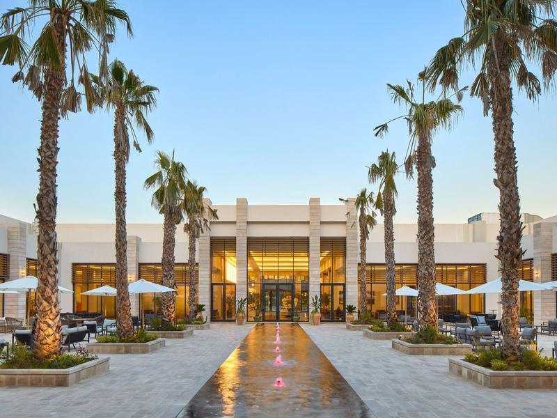 Moderner Innenhof mit Palmen, Sitzbereichen und beleuchtetem Eingang bei klarem Himmel.