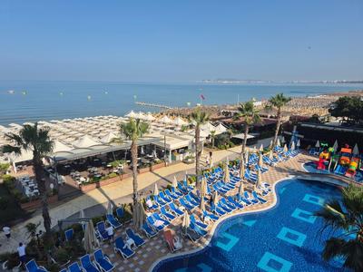 Hotel pool with blue loungers, palm trees, and beach view under clear sky.
