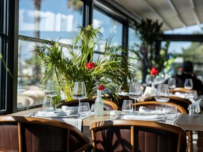 Elegantes Restaurant mit gedeckten Tischen und grüner Dekoration neben großen Fenstern.
