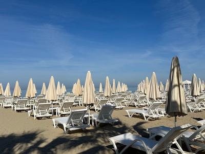 Sandstrand mit vielen weißen Liegestühlen und geschlossenen Sonnenschirmen unter blauem Himmel.