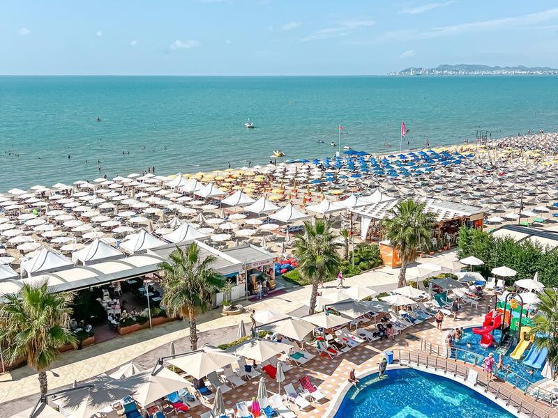 View of a pool with blue tile pattern and a beach with many umbrellas by the sea.