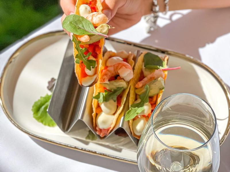 Hand holding a plate with three tacos and a glass of white wine on a white table.
