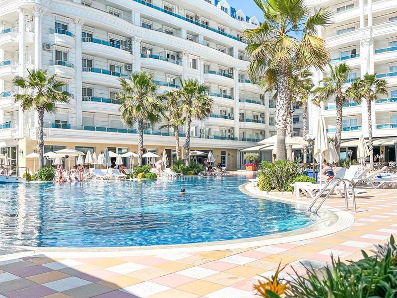 Modern hotel complex with large swimming pool, palm trees, and blooming flowers in the foreground.