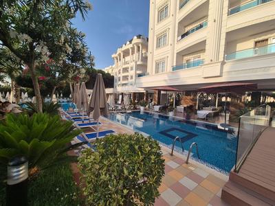 Hotelpool mit Liegestühlen, Sonnenschirmen und angrenzendem Garten unter blauem Himmel.
