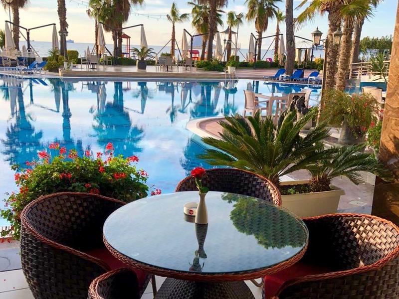 Pool area with round tables and chairs, surrounded by palm trees and sea view.