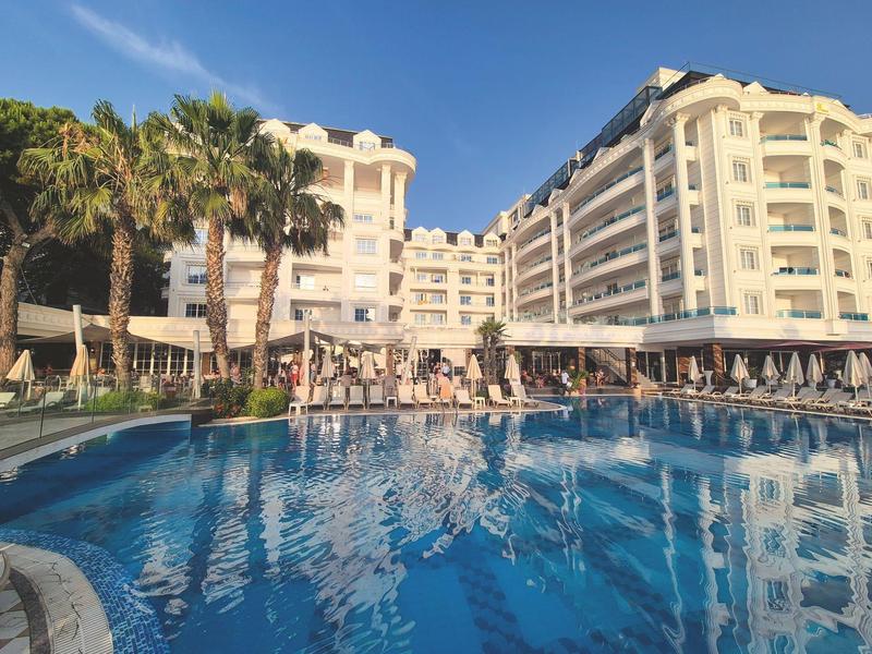 Large hotel building with pools, palm trees, and umbrellas under clear sky.