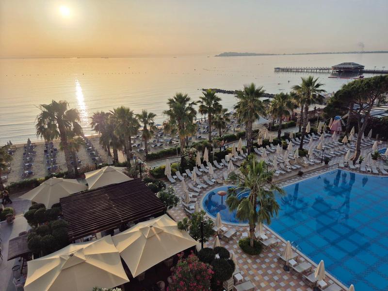 View of a hotel pool with sun loungers and palm trees by the sea at sunset.