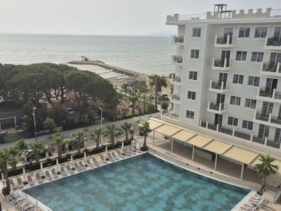 Hotelpool mit Liegestühlen neben einem weißen Gebäude und Blick auf das Meer und eine Grünfläche.