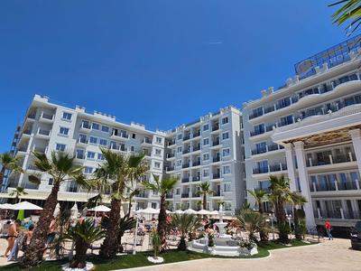 Großes weißes Hotelgebäude mit mehreren Etagen, Palmen und Sonnenschirmen im Außenbereich.