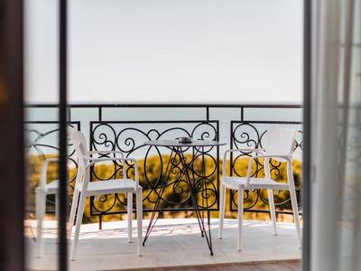 Zwei weiße Stühle und ein Glas auf einem Balkon mit kunstvollem Geländer und Blick aufs Meer.