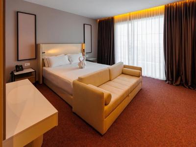 Modern eingerichtetes Hotelzimmer mit Doppelbett, Sofa und großem Fenster mit Vorhängen.