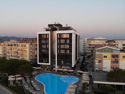 Moderner Hotelkomplex mit geschwungenem Pool und Sonnenliegen in urbaner Umgebung bei Dämmerung.