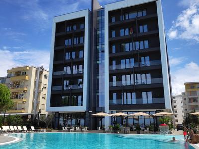 Moderner Hotelkomplex mit großem Außenpool vor einem mehrstöckigen Gebäude unter blauem Himmel.