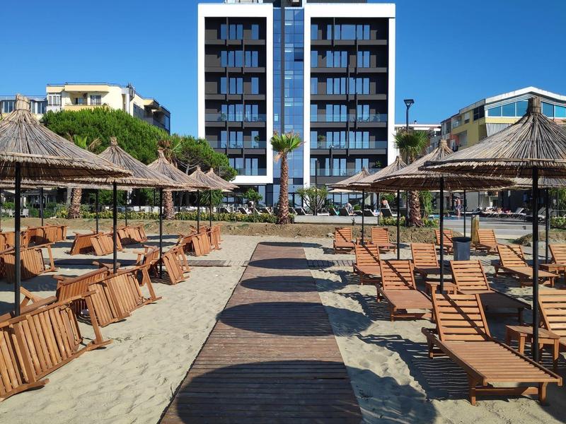 Sicht auf einen Sandstrand mit Holzliegen und Strohdächern vor einem modernen Hotel bei klarem Himmel.