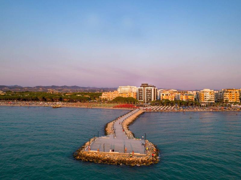 Mole mit Steinen und Beton führt von Wasser zu einem belebten Küstenort bei klarem Himmel.