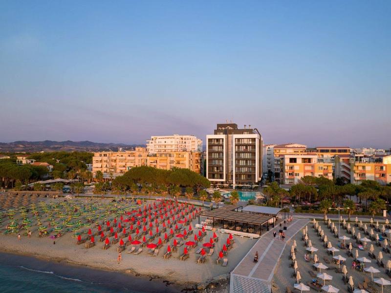 Strand mit Reihen farbiger Sonnenschirme und dahinter mehrstöckige Gebäude bei klarem Himmel.