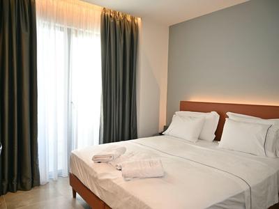 Hell eingerichtetes Schlafzimmer mit Doppelbett, weißen Bettdecken und dunklen Vorhängen am Fenster.