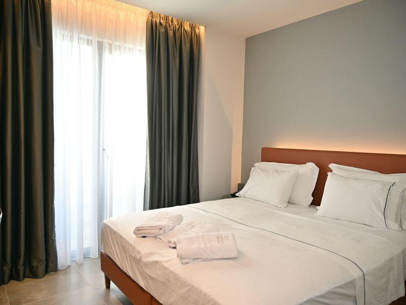 Jasná ložnice s manželskou postelí a velkým oknem s závěsy.