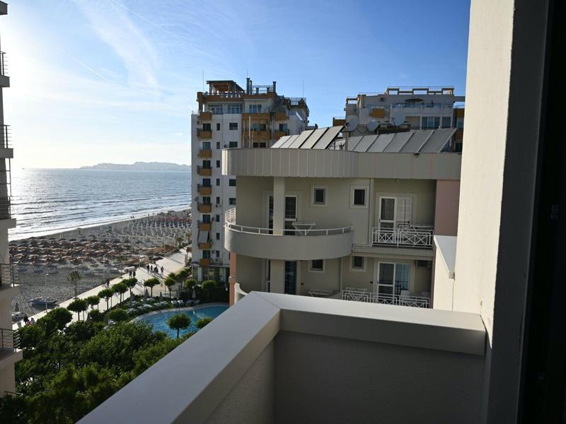 Výhled z balkonu na sousední budovy, bazén a pláž u moře.