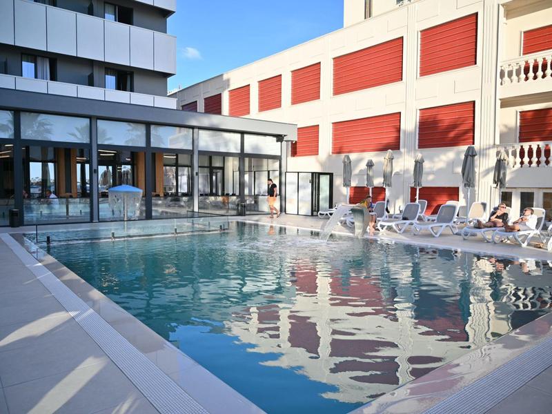 Moderní venkovní bazén s lehátky a okolními hotelovými budovami pod jasnou oblohou.