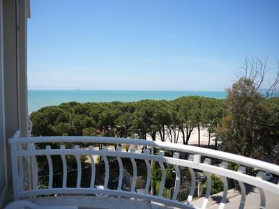 Balkon mit weißem Geländer, Blick auf grüne Bäume und Meer unter klarem Himmel.