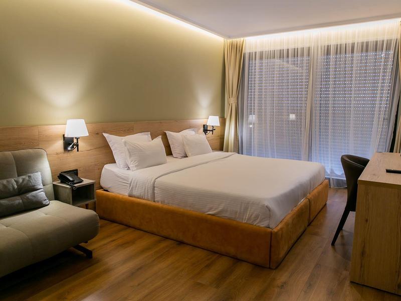 Modern hotel room with a cozy double bed, sofa, desk, and large windows with curtains.