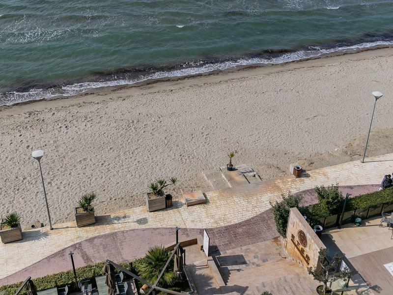 View of a sandy beach with walkway, lamps, and plant pots next to a hotel area.