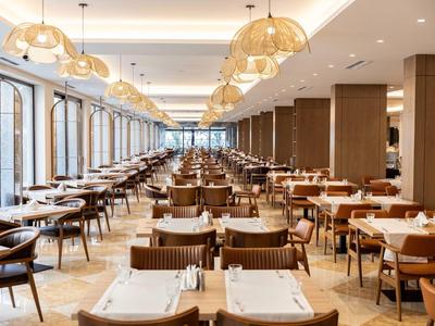 Großer, moderner Hotel-Speisesaal mit Holzstühlen und Tischen, großen Fenstern und Hängeleuchten.