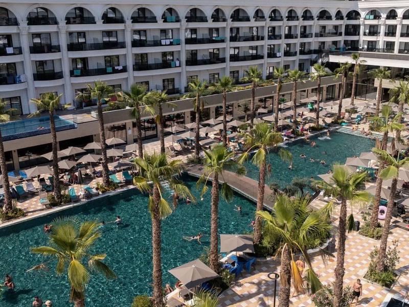 Großer Hotelpool mit Palmen, Liegestühlen und mehrstöckigem Gebäude im Hintergrund.