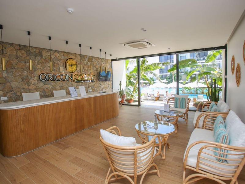 Heller Hotel-Empfang mit Holztheke und Korbmöbeln vor großen Fenstern.