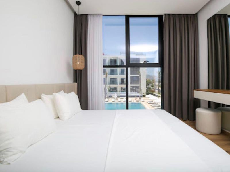 Modernes Hotelzimmer mit großem Bett, Kissen und Blick auf Pool und Gebäude durch Fenster.