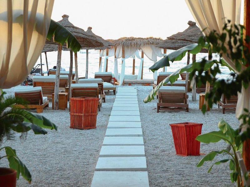 Blick durch Vorhänge auf einen Weg mit Liegestühlen und Sonnenschirmen am Strand.