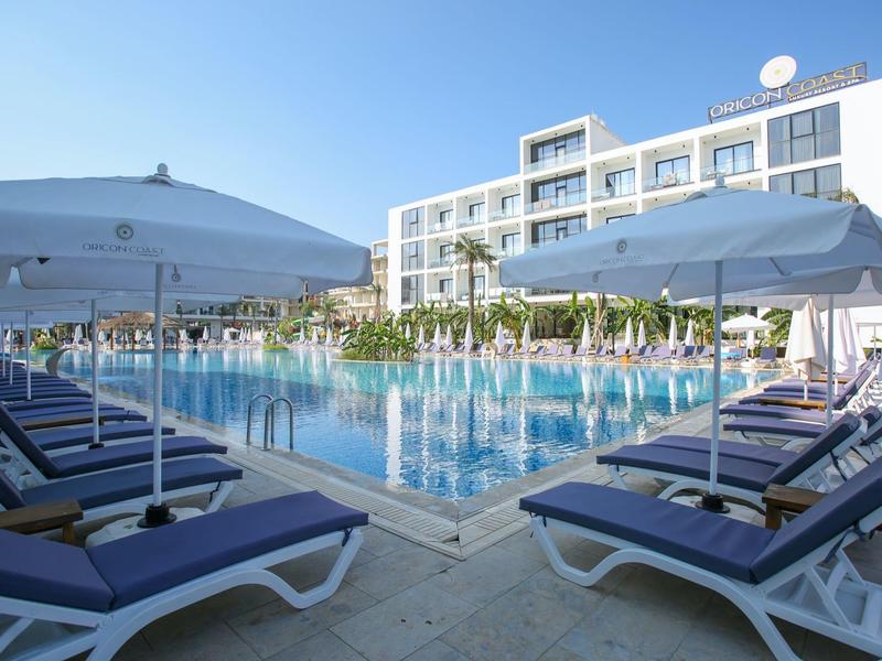 Schwimmbad mit blauen Liegestühlen und Sonnenschirmen vor einem modernen Hotelgebäude bei klarem Himmel.