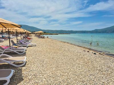 Strand mit Liegestühlen und Sonnenschirmen neben klarem Meer und Bergen im Hintergrund.