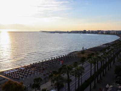 Sonnenuntergang über einem Strand mit Sonnenschirmen und Palmen neben einer Promenade.