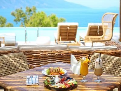 Terrasse mit Tisch, zwei Getränken und Speisen, Blick auf Pool und Meer im Hintergrund.