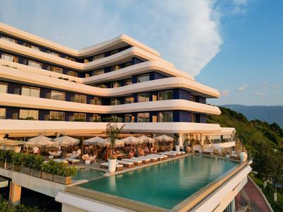 Moderner mehrstöckiger Hotelkomplex mit Terrasse, Pool und Sitzbereichen, umgeben von grüner Landschaft.